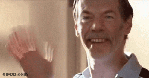 A Man With A Beard Is Smiling And Waving His Hand In Front Of A Window . GIF