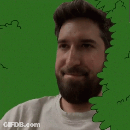 A Man With A Beard Is Smiling Behind A Green Bush GIF