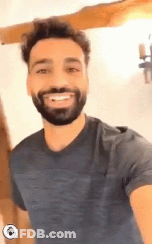 A Man With A Beard Is Smiling For The Camera While Wearing A Blue Shirt . GIF
