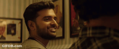 A Man With A Beard Is Smiling In Front Of A Wall With Pictures On It GIF