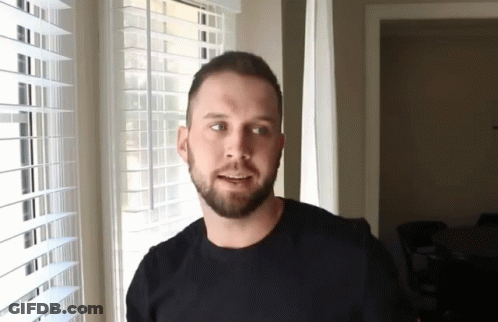 A Man With A Beard Is Smiling In Front Of A Window With Blinds GIF