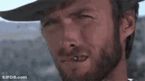 A Man With A Beard Is Smoking A Cigarette And Wearing A Cowboy Hat . GIF