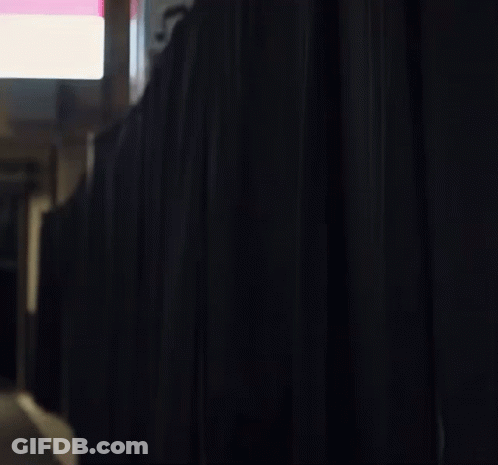 A Man With A Beard Is Standing Behind A Black Curtain In A Room . GIF