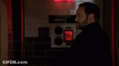 A Man With A Beard Is Standing In A Dark Room With A Red Light Behind Him . GIF