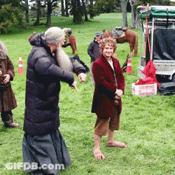 A Man With A Beard Is Standing In A Field With Other People And A Horse In The Background GIF