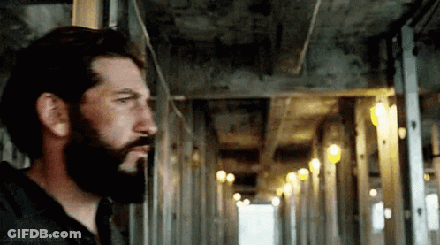 A Man With A Beard Is Standing In A Hallway Looking Out The Window . GIF