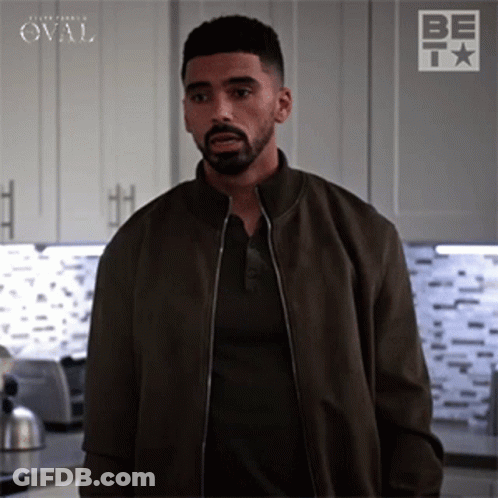 A Man With A Beard Is Standing In A Kitchen Wearing A Brown Jacket And A Black Shirt . GIF