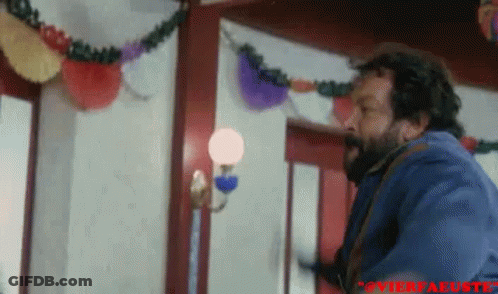 A Man With A Beard Is Standing In A Room With A Lamp And Decorations On The Wall . GIF