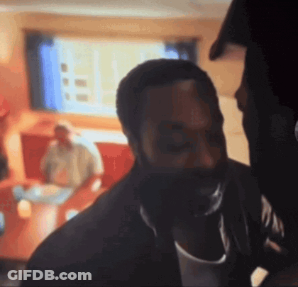 A Man With A Beard Is Standing In A Room With A Man Sitting On A Couch Behind Him . GIF