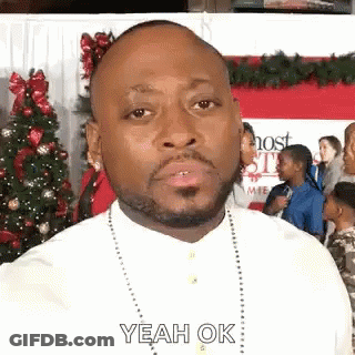 A Man With A Beard Is Standing In Front Of A Christmas Tree And Making A Funny Face . GIF
