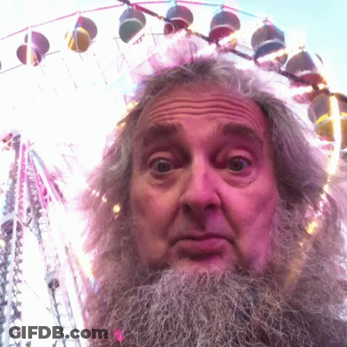 A Man With A Beard Is Standing In Front Of A Colorful Ferris Wheel GIF