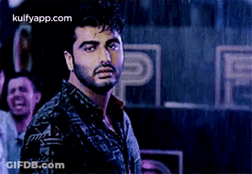 A Man With A Beard Is Standing In Front Of A Crowd Of People In A Dark Room . GIF