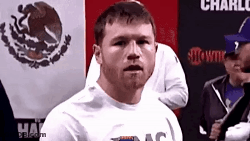 A Man With A Beard Is Standing In Front Of A Mexican Flag And Looking At The Camera . GIF