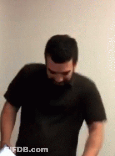 A Man With A Beard Is Standing In Front Of A White Wall Wearing A Black Shirt GIF