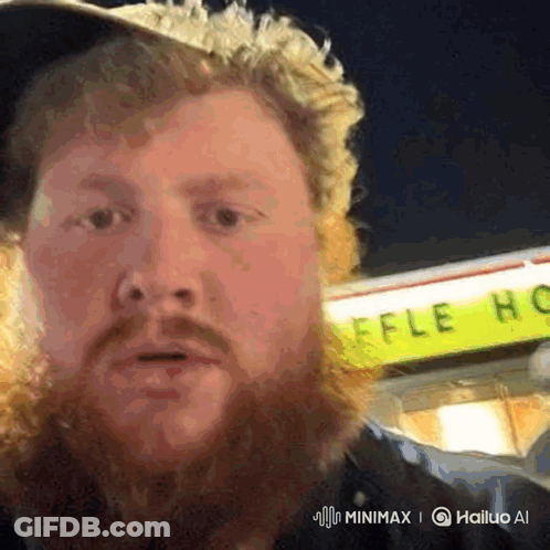 A Man With A Beard Is Standing In Front Of A Yellow Sign That Says Waffle House GIF
