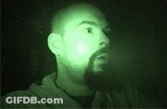 A Man With A Beard Is Standing In The Dark With A Green Light Shining On His Face . GIF
