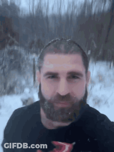 A Man With A Beard Is Standing In The Snow Wearing A Black Shirt With The Letter T On It GIF