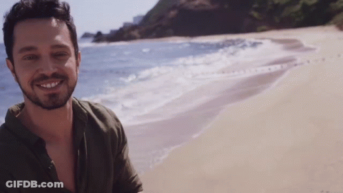 A Man With A Beard Is Standing On A Beach And Smiling GIF