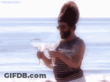 A Man With A Beard Is Standing On A Beach Holding A Martini Glass . GIF