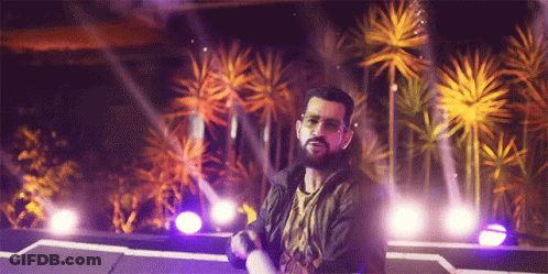 A Man With A Beard Is Standing On A Stage In Front Of Palm Trees And Purple Lights GIF