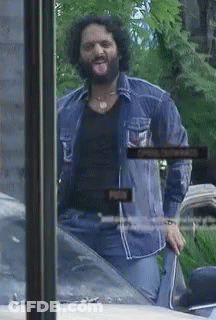 A Man With A Beard Is Sticking His Tongue Out In Front Of A Sign That Says Open GIF