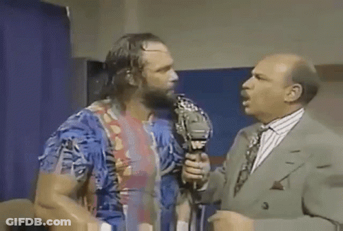 A Man With A Beard Is Talking Into A Microphone While Another Man In A Suit Stands Behind Him . GIF
