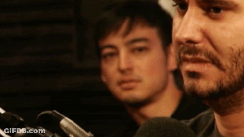A Man With A Beard Is Talking Into A Microphone While Another Man Watches . GIF