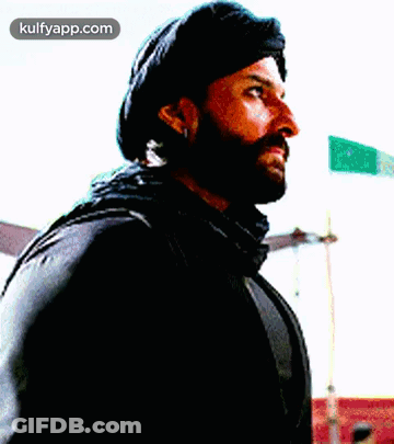 A Man With A Beard Is Wearing A Black Hat And Scarf And Looking To The Side . GIF