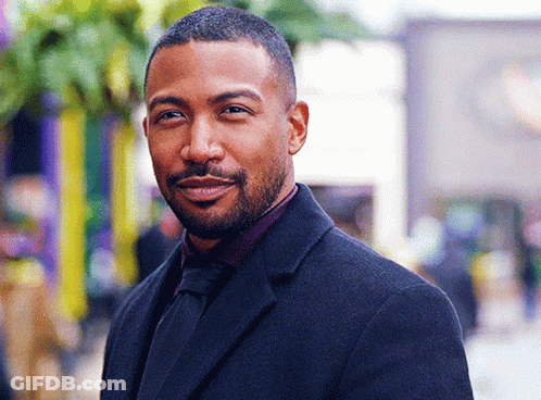 A Man With A Beard Is Wearing A Black Suit And Tie And Smiling . GIF