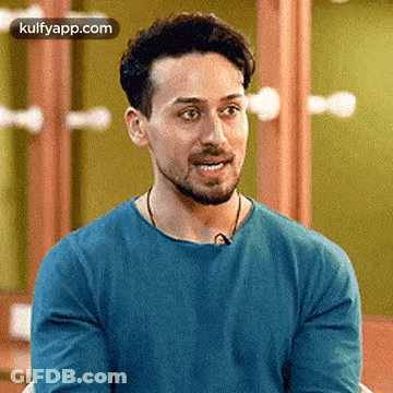 A Man With A Beard Is Wearing A Blue Shirt And Smiling While Sitting In Front Of A Mirror . GIF