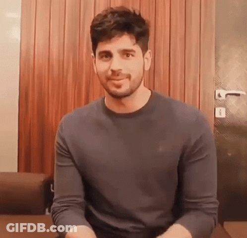 A Man With A Beard Is Wearing A Grey Sweater And Smiling While Sitting On A Couch . GIF