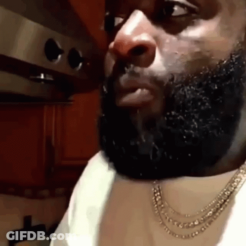 A Man With A Beard Is Wearing A Necklace And A White Shirt In A Kitchen . GIF