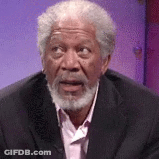 A Man With A Beard Is Wearing A Suit And A Pink Shirt And Making A Surprised Face . GIF
