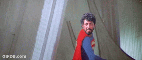 A Man With A Beard Is Wearing A Superman Costume And Standing In Front Of A Wall . GIF