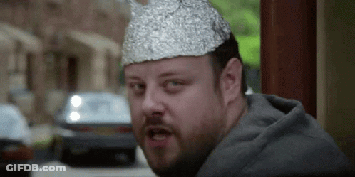 A Man With A Beard Is Wearing A Tin Foil Hat And Making A Funny Face . GIF