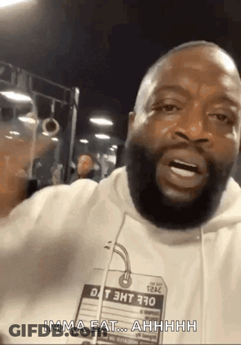 A Man With A Beard Is Wearing A White Hoodie With A Tag That Says ' Imma Eat ' On It . GIF