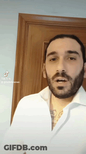 A Man With A Beard Is Wearing A White Shirt And A Tattoo On His Chest . GIF