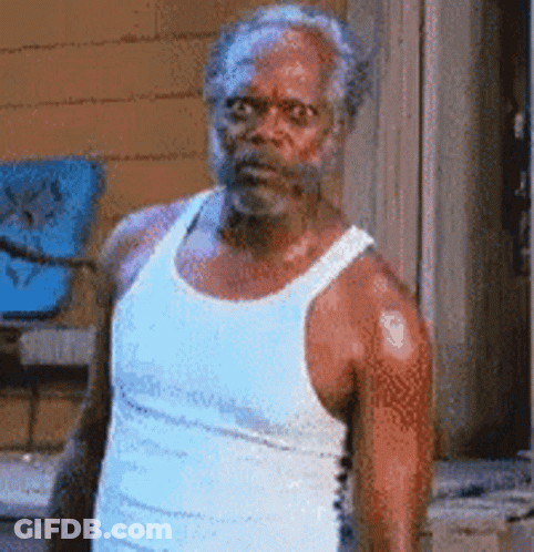 A Man With A Beard Is Wearing A White Tank Top And Standing In Front Of A Building . GIF