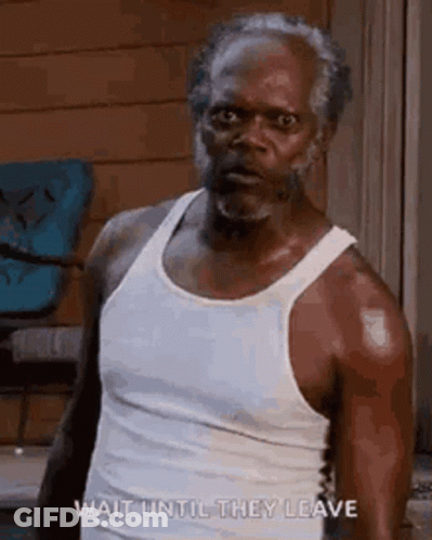 A Man With A Beard Is Wearing A White Tank Top And Standing In Front Of A Chair . GIF