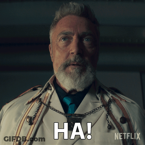 A Man With A Beard Is Wearing A White Trench Coat And A Necklace With The Word Ha On It GIF