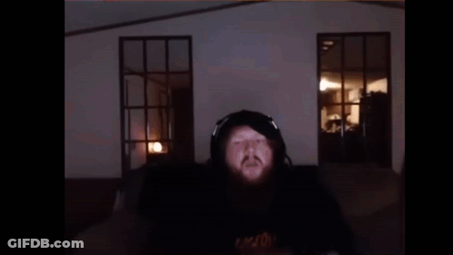 A Man With A Beard Is Wearing Headphones And Making A Funny Face While Sitting In Front Of A Window . GIF