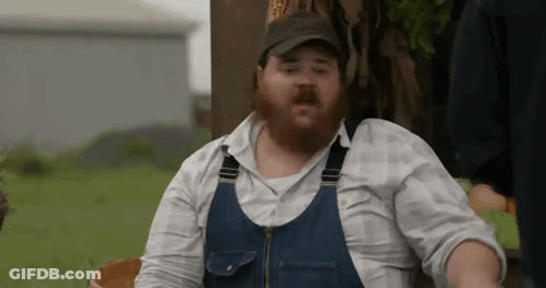 A Man With A Beard Is Wearing Overalls And A Baseball Cap . GIF