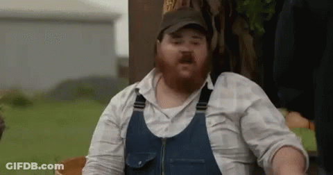 A Man With A Beard Is Wearing Overalls And A Hat While Sitting In A Field . GIF