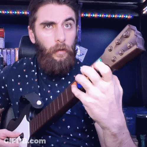 A Man With A Beard Playing A Guitar In Front Of A Bookshelf GIF