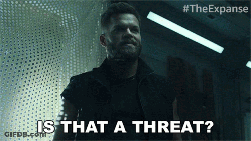A Man With A Beard Says " Is That A Threat " In A Dark Room GIF