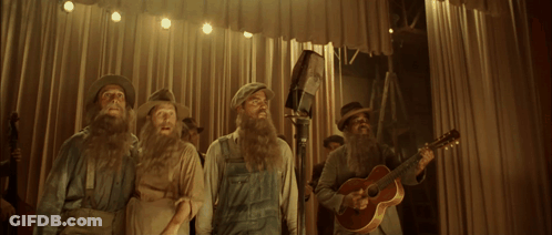 A Man With A Beard Sings Into A Microphone While Other Men Play Instruments GIF