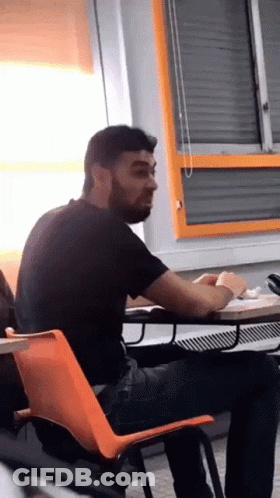 A Man With A Beard Sits At A Desk In A Classroom GIF