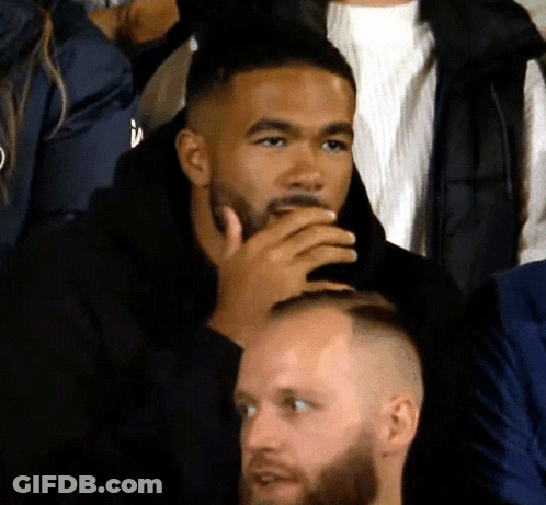 A Man With A Beard Sits In A Crowd With His Hand To His Mouth GIF