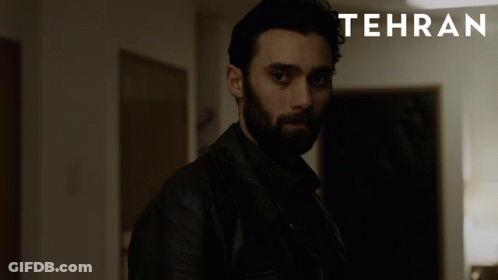 A Man With A Beard Stands In A Dark Room With Tehran Written On The Bottom GIF