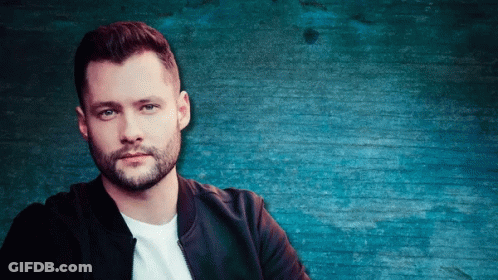 A Man With A Beard Stands In Front Of A Blue Brick Wall GIF